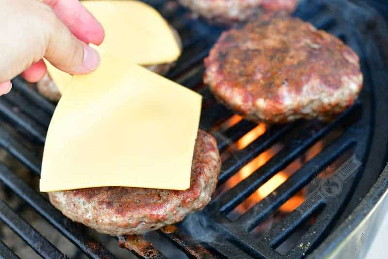 placing a slice of American cheeseon a cooked burger patty on the grill