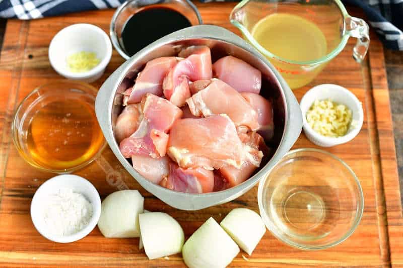 ingredients in bowls on a wooden cutting board including cut chicken meat, onions, corn starch, garlic, vinegar, honey, broth, soy sauce, and ginger