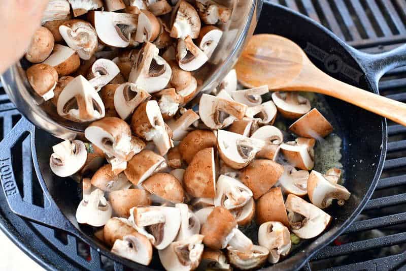 pouring in cut mushrooms into the cast iron skillet on the grill