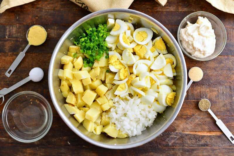 chopped potatoes, eggs, onion, and green onion in a metal bowl with a small bowl of mayo and vinegar next to it and measuring spoons of mustard, salt, pepper, and garlic powder next to the mixing bowl