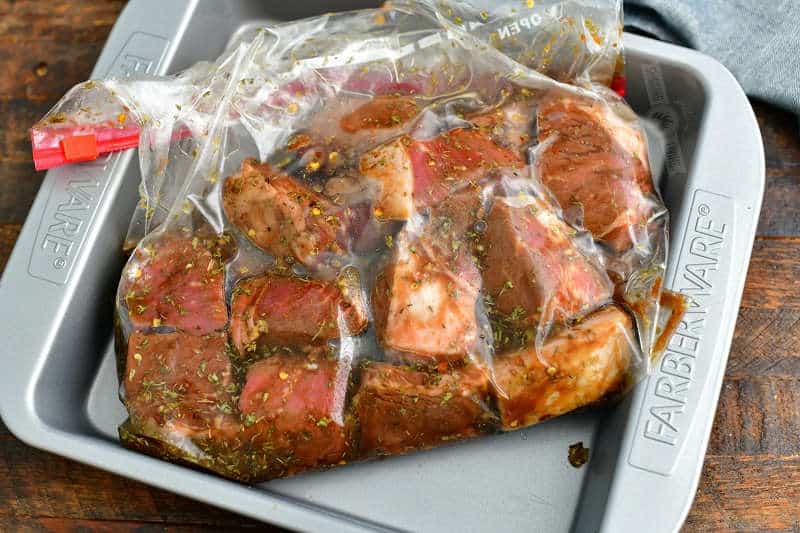 cubed steak pieces in marinade inside a plastic sip-top bag placed in a metal square pan