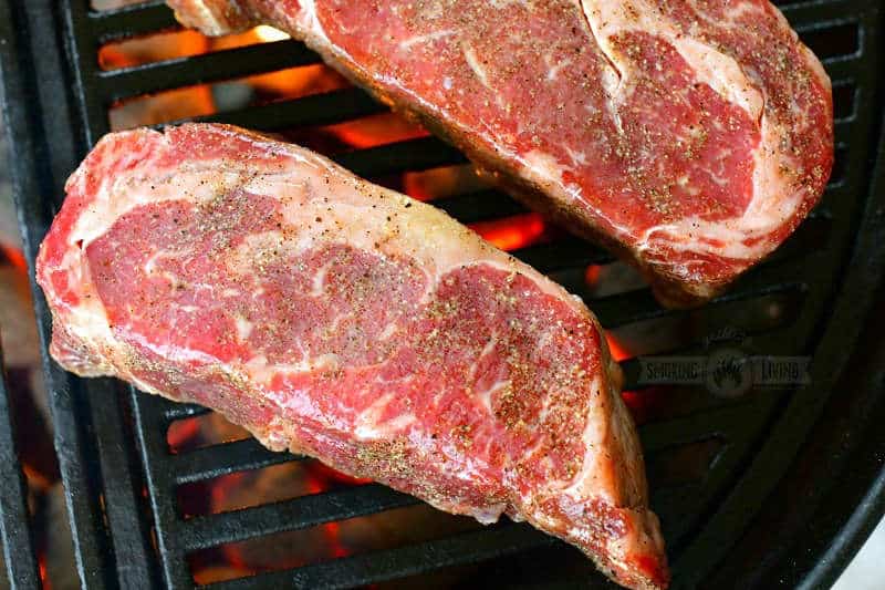 raw steaks seasoned with salt and pepper on the grill with hot coals visible below