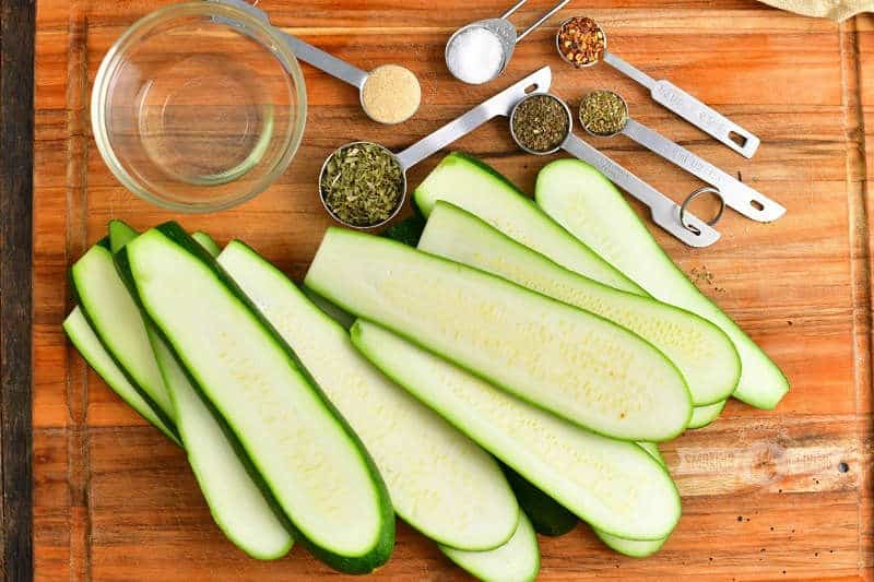 sliced raw zucchini, oil in a small bowl, measuring spoons with parsley, basil, oregano, salt, garlic powder, and red pepper flakes on a wooden cutting board