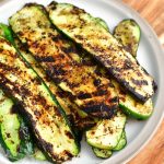 top view of grilled zucchini slices on a plate on to of a wooden cutting board with some brown towel visible