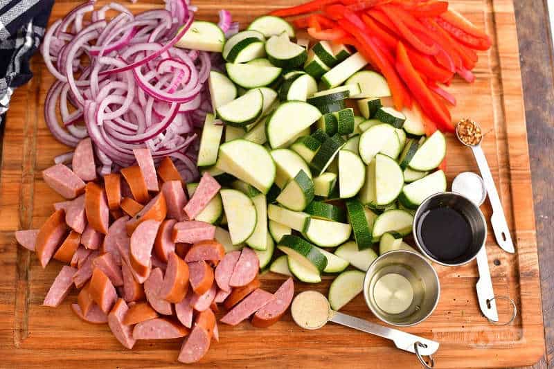 diced zucchini, smoked sausage, sliced bell pepper, sliced onion, oil and vinegar in silver cups and seasonings in measuring spoons on a wooden cutting board