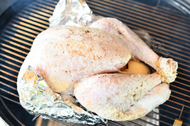 uncooked turkey on the smoker side view with wings wrapped in foil