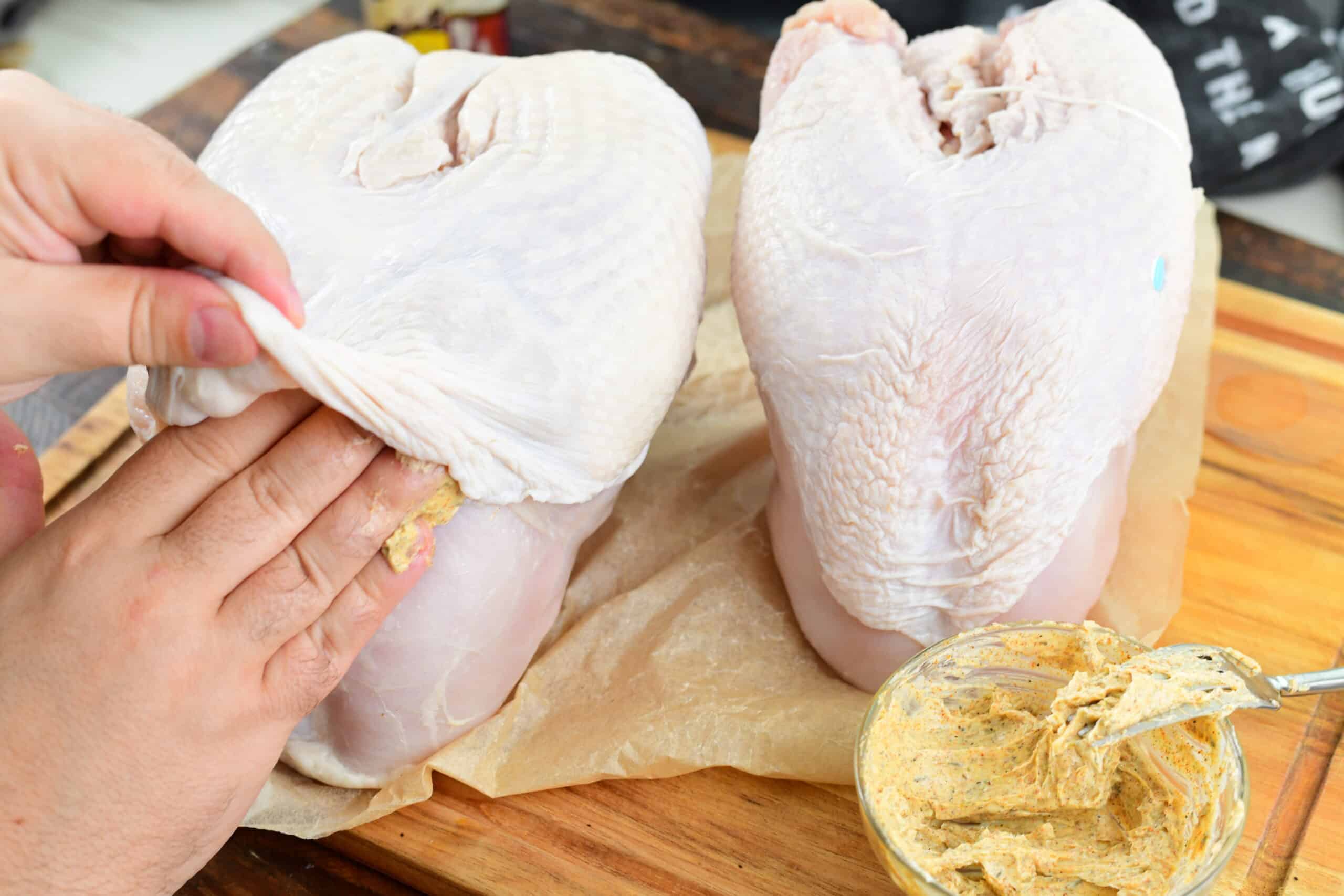 raw turkey on a cutting board with seasoned butter in a glass bowl with a fork and a hand rubbing the turkey under the skin with seasoned butter.