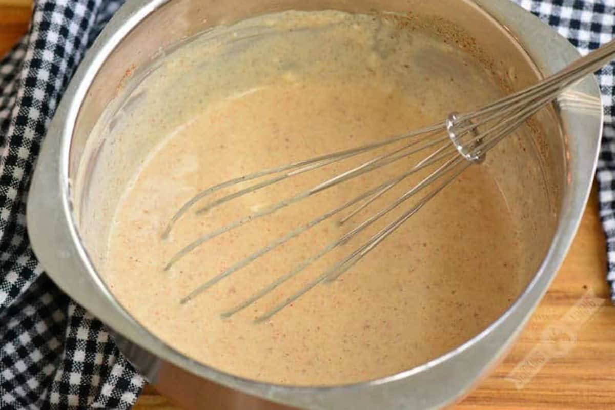 White BBQ sauce in a metal bowl with a metal whisk on a wood surface.